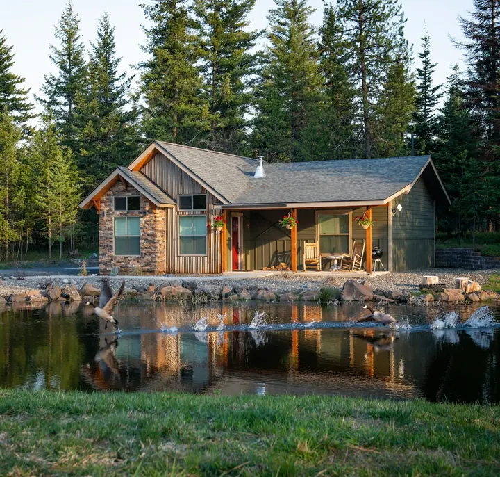 Stone-and-wood lake cottage among tall pines, reflected in calm water at sunset